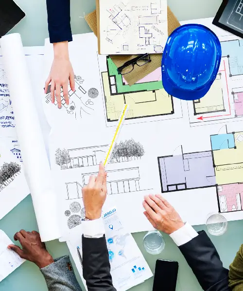 Top-down view of hands pointing at architectural blueprints and colored floor plan drawings with a blue safety helmet on the table